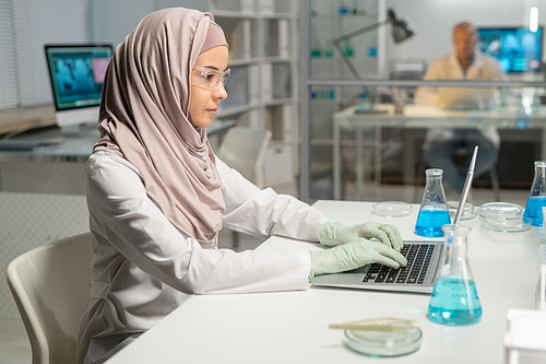 Young Muslim woman in hijab, lab coat, protective eyeglasses and gloves sitting by desk in front of laptop in laboratory and typing