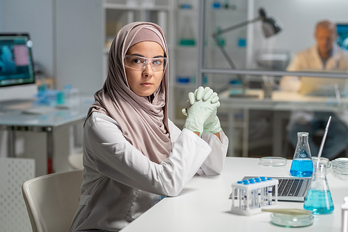 Young contemporary female researcher in protective eyeglasses and hijab looking at camera while sitting by desk in front of laptop
