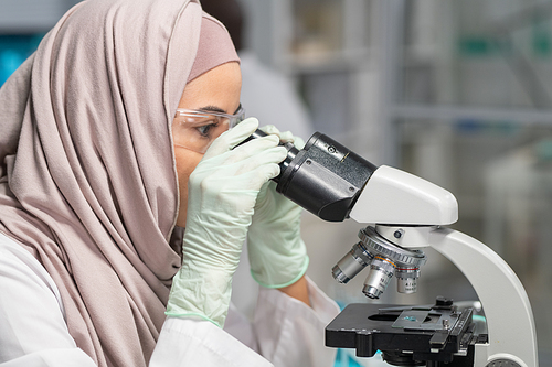 Side view of young Muslim female scientist in hijab, gloves and protective eyewear studying new chemical compound in laboratory