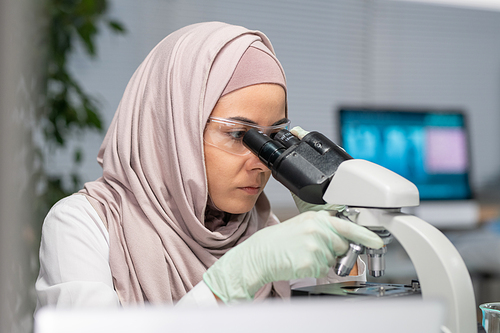 Young serious Muslim woman in gloves, protective eyeglasses and hijab studying characteristics of chemical substance in microscope