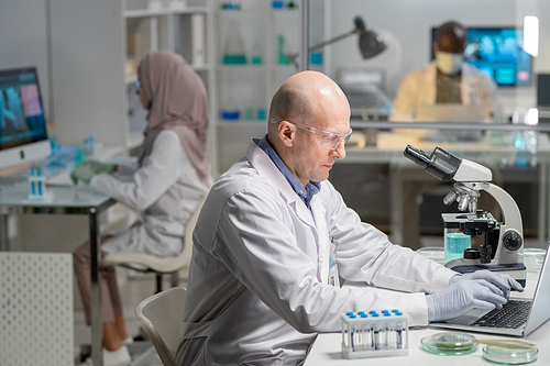 Mature serious male lab worker in gloves, whitecoat and protective eyeglasses searching for scientific information in the net