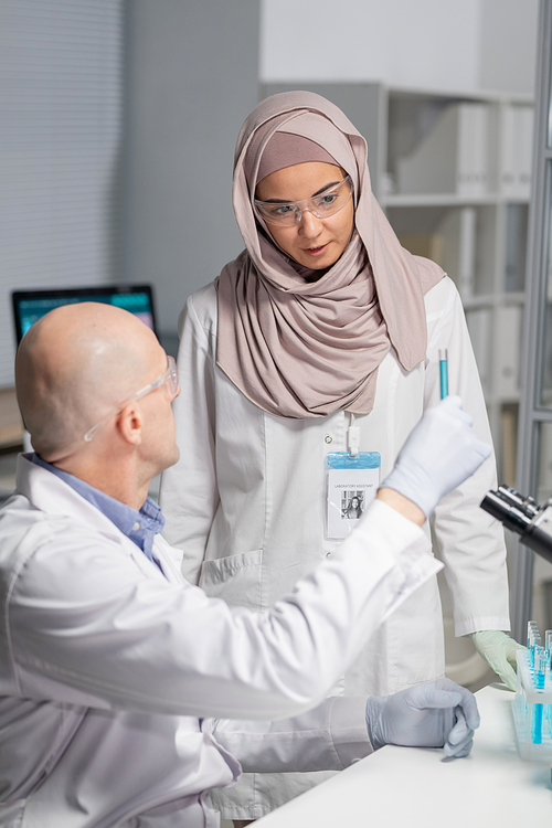 Young Muslim female virologist in hijab and whitecoat looking at blue liquid in flask held by mature male colleague in laboratory