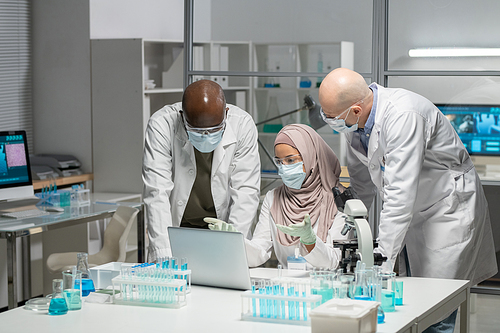 Young Muslim female researcher in hijab, whitecoat, protective gloves, mask and eyeglasses making presentation to male colleagues