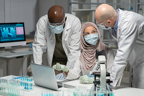 Three interracial colleagues in protective workwear having discussion of results of clinical or scientific experiment in laboratory