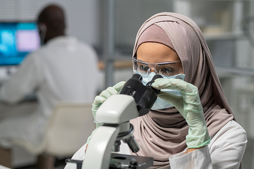 Contemporary female researcher in hijab looking in microscope while studying new chemical substances or bacteria in laboratory
