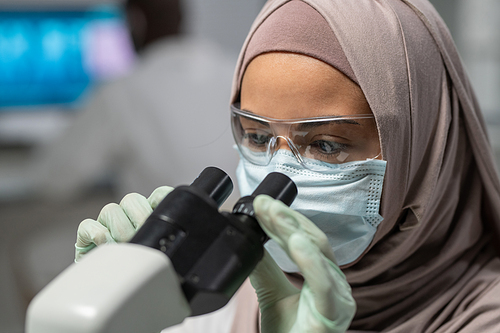Close-up of young Muslim female researcher looking in microscope during scientific investigation or discovery of new virus