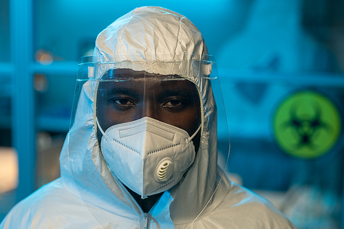 Shot of young contemporary African American scientist in coveralls, protective screen on face and respirator looking at camera