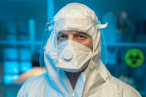 Face of male researcher in biohazard suit, respirator and protective screen looking at camera against interior of laboratory