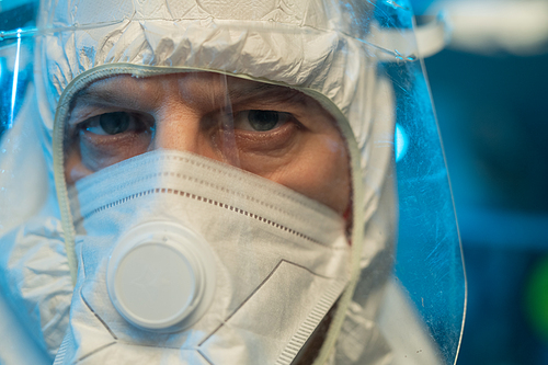 Mature male virologist or researcher in white biohazard suit, respirator and protective facial screen looking at camera