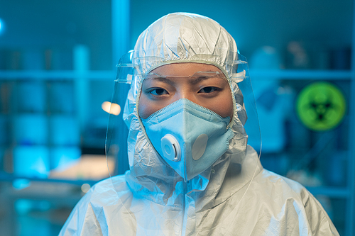 Young Asian female microbiologist or clinician in protective coveralls looking at camera in modern scientific laboratory