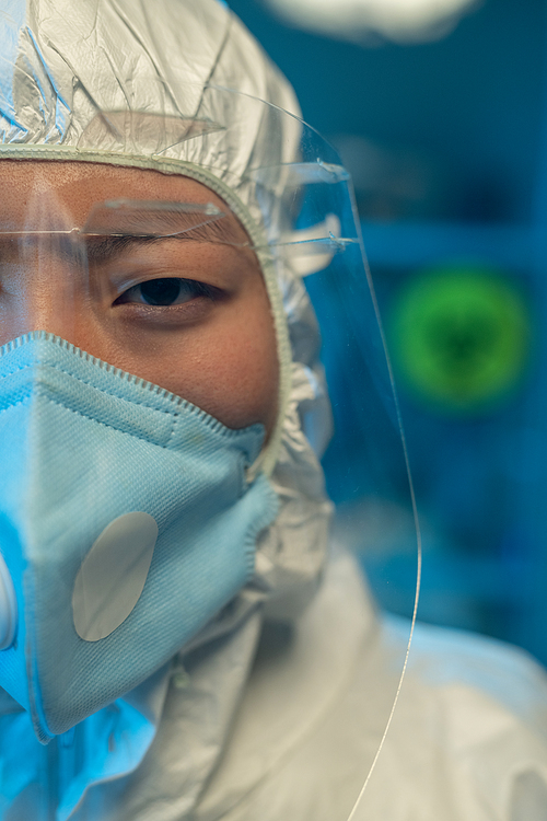Half of face of young Asian woman in coveralls, respirator and protective eyeglasses standing in front of camera in scintific laboratory