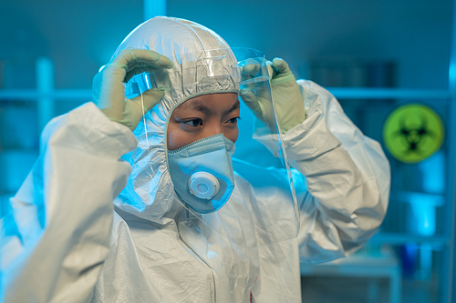 Young Asian female scientist in biohazard suit, gloves and respirator putting protective screen on her face in front of laboratory