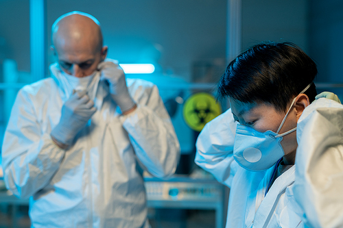 Young Asian female and her male colleague in white coveralls putting on respirators while going to enter scientific laboratory
