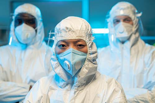 Three interracial scientists in protective coveralls, respirators and screens standing in front of camera against chemical laboratory
