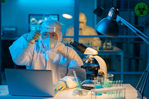 Young female virologist in protective coveralls, respirator, gloves and facial screen studying sample of new vaccine in laboratory