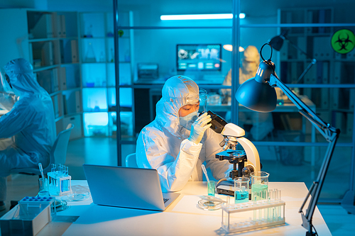 Female scientist in biohazard suit, respirator and gloves using microscope for studying new substances and their characteristics