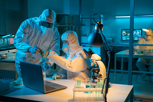 Two young chemists in protective coveralls, gloves and respirators studying liquid substance in flask by workplace in laboratory