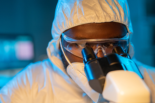 Close-up of young African American scientist in protective coveralls, respirator and eyeglasses looking in microscope in laboratory