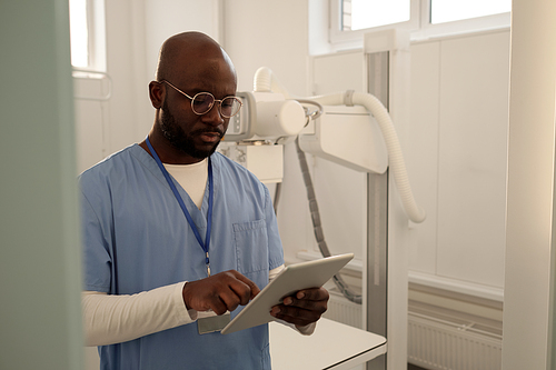 Young African American clinician in medical scrubs and eyeglasses using tablet while looking through online data or electronic form