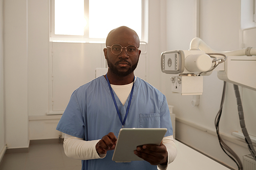 Young serious African American male clinician in uniform with tablet looking at camera while consulting patients online