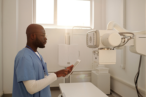 African American male clinician choosing settings of x-ray machine in tablet while standing in front of medical equipment