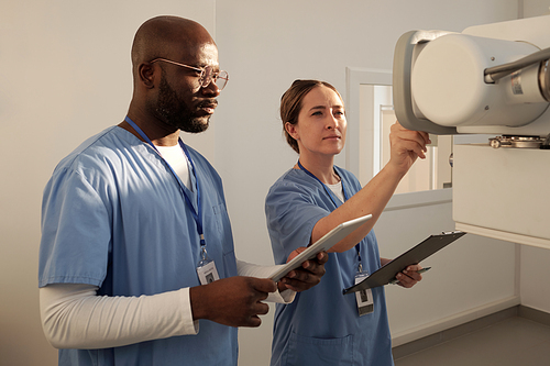 Two young interracial clincians in uniform switching on medical equipment while female pressing button on tableau