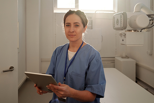 Young pretty female radiologist with tablet standing in front of camera against x-ray equipment while working in hospital