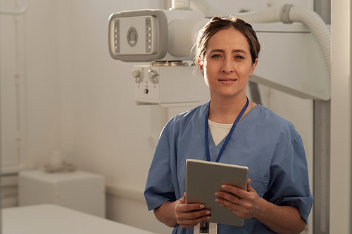 Happy young contemporary female clinician in blue uniform holding tablet while standing against medical equipment
