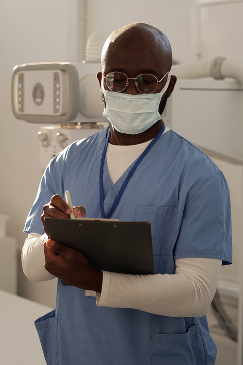 Young serious African American clinician in uniform and protective mask making notes or prescriptions for patient in hospital