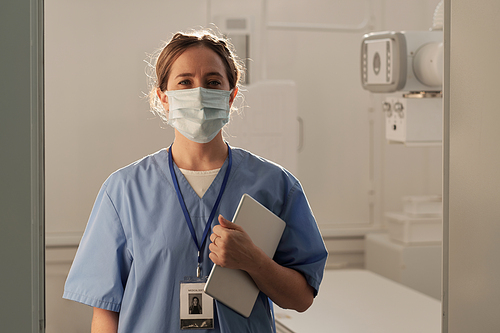 Pretty female healthcare worker in blue uniform and protective mask standing in medical office with x-ray equipment on background
