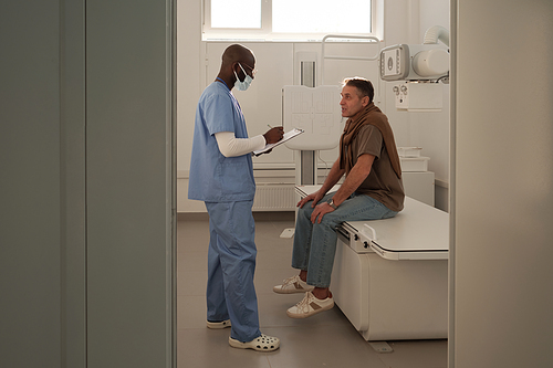 Young doctor communicating with mature male patient before x-ray examination and making notes in medical form