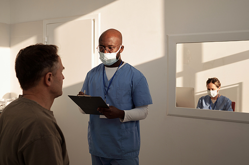 Mature male patient describing his symptoms to young physician in uniform and protective mask making notes in document