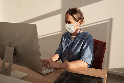 Young serious female clinician in mask and uniform entering medical data in computer while sitting by desk in front of monitor