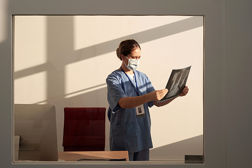 Young female radiologist in uniform and protective mask looking at x-ray image of lings of patient while standing by workplace