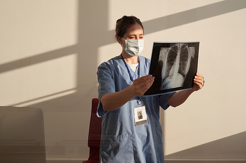 Young serious female radiologist looking at x-ray image after radiological examination of patient while standing in medical office