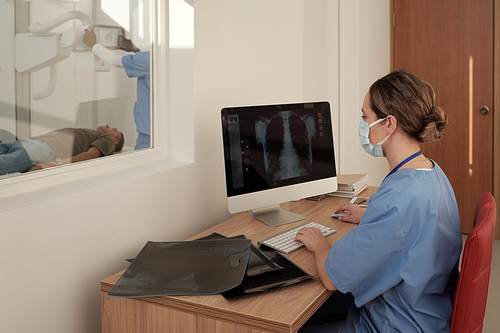 Young female radiologist in mask sitting by workplace in front of computer and looking at x-ray image of patient lungs or thorax