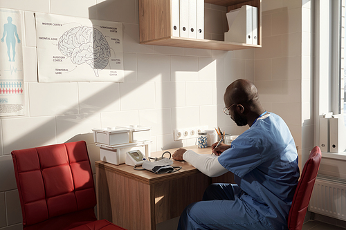 Young African American clinician making medical notes in document by his workplace in office while waiting for new patients