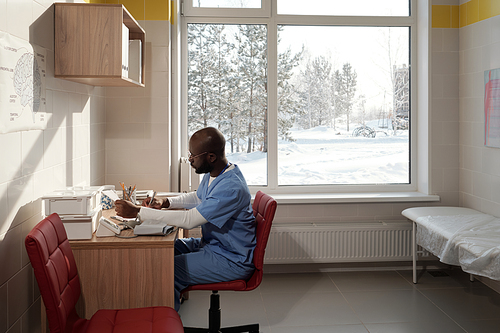 Young contemporary black man in medical scrubs making prescription notes in card of patient while sitting by desk against window