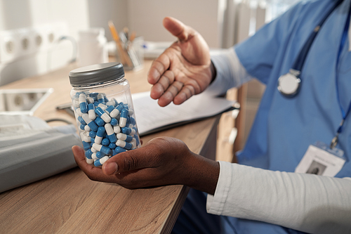Hands of young African American male clinician holding jar with white and blue pills while recommending them to patient