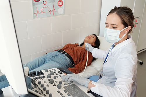Female endocrinologist in whitecoat and mask sitting by ultrasound equipment during examination of Hispanic patient on couch