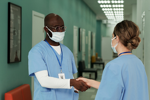 Young interracial healthcare workers shaking hands of one another at the beginning of new working day in hospital corridor