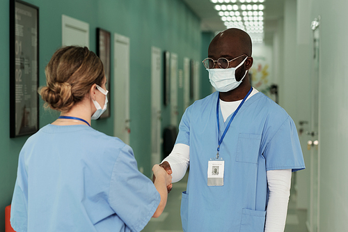 Young African American surgeon in uniform and protective mask shaking hand of his female assistant while both standing in hospital corridor
