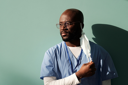 Tired African American doctor in blue uniform putting off protective mask after hard working day while standing against wall