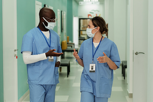 Two young intercultural doctors or surgeons in protective masks having discussion of troubles while walking along hospital corridor
