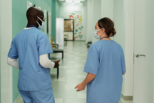Rear view of two young interracial clinicians in blue uniform and protective masks having discussion in hospital corridor