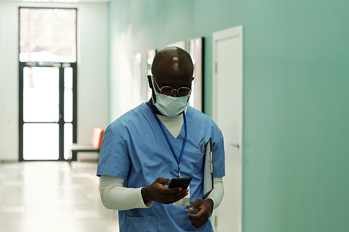 Young black man in blue uniform and protective masks texting in smartphone while moving along corridor after work