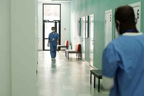 Young female clinician in blue uniform and protective mask walking along hospital corridor towards African American doctor