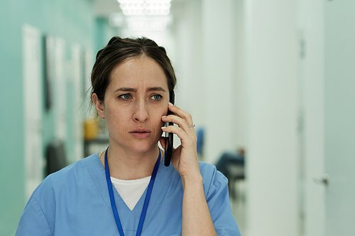 Young troubled female clinician or assistant in blue uniform speaking on mobile phone while moving along in hospital corridor