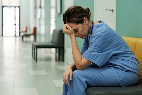 Young tired or stressed female doctor in uniform sitting in hospital corridor with her head in hands after shocking news