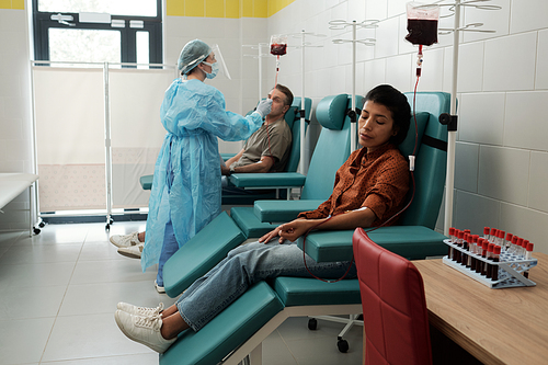 Young Hispanic female donor giving her blood for sick patients of hospital while sitting in armchair in large ward
